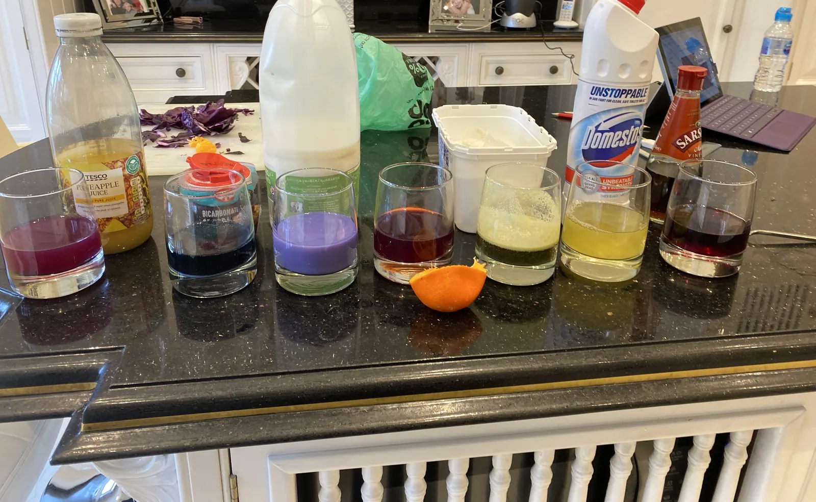 A row of glasses holding red cabbage indicator, each turned a different colour by household liquids