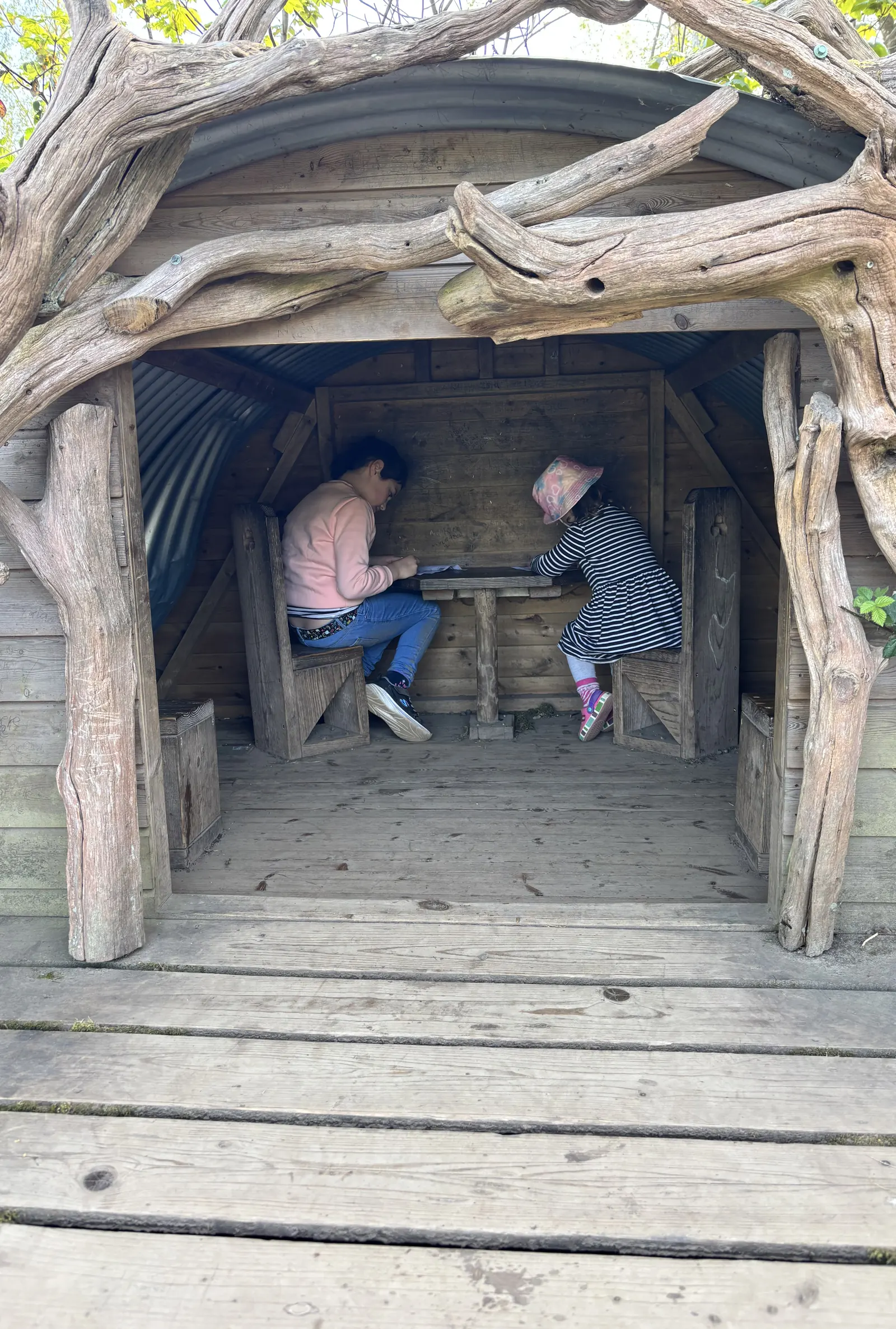 Two children reading together inside a wooden den in a garden